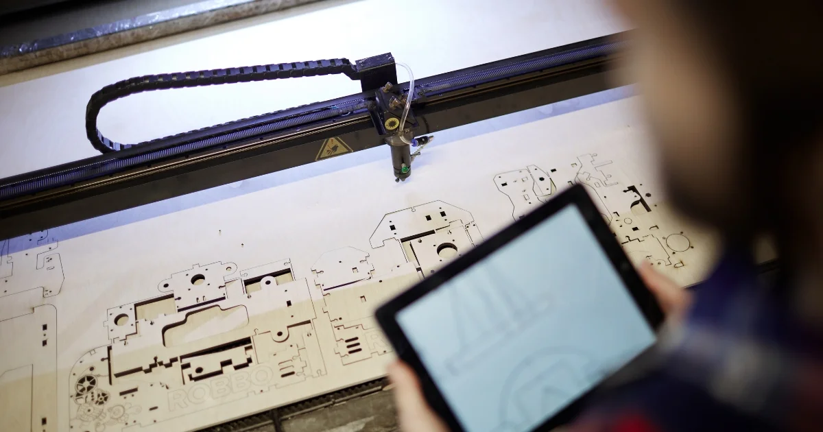 A laser cutting machine engraves intricate designs on a wooden sheet, while an operator monitors progress on a tablet.